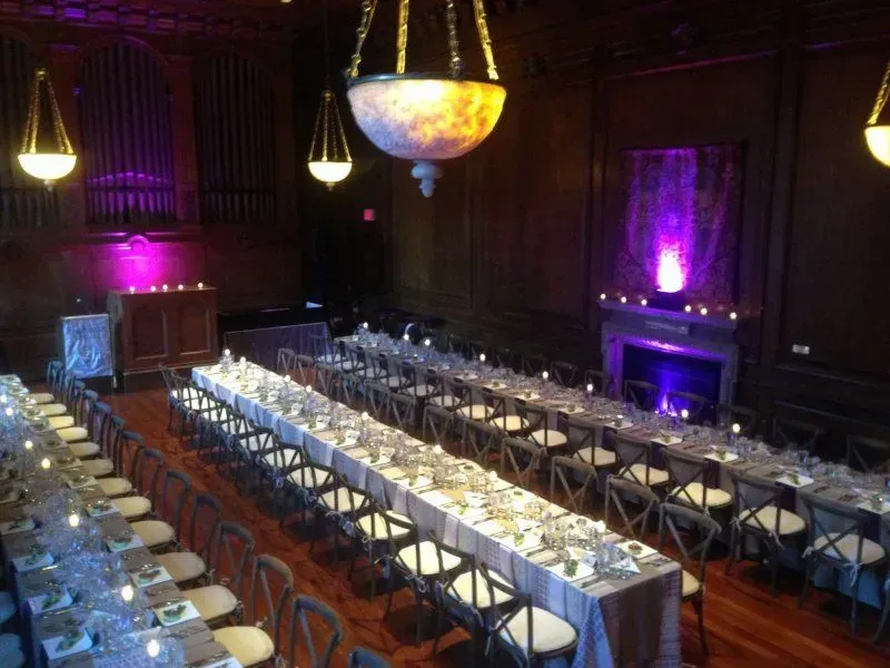 A banquet hall with long tables set for dinner, featuring elegant hanging lights and purple accent lighting.