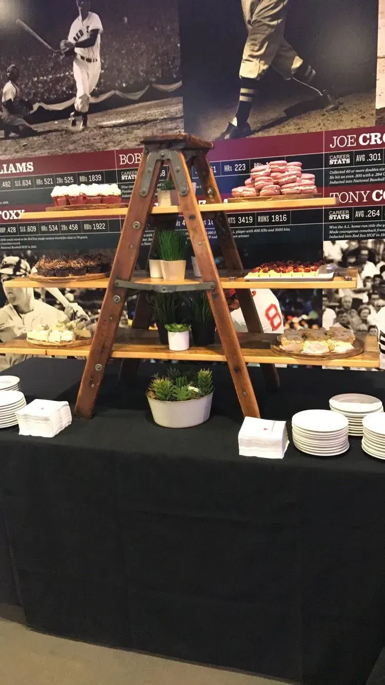 A tiered dessert display made from a wooden ladder on a black-clothed table in front of a baseball-themed wall mural.