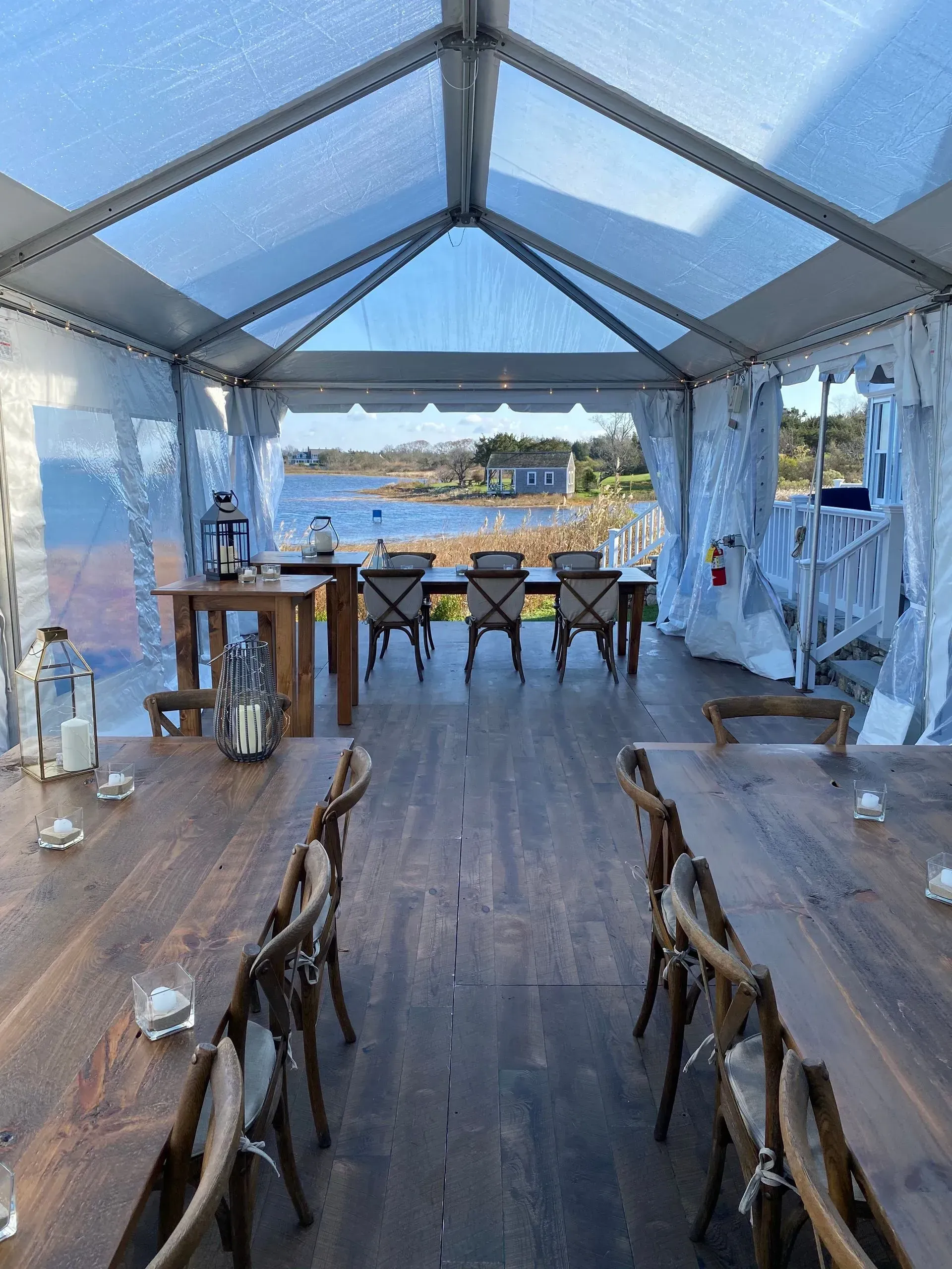 A glass-walled tent interior with wooden tables and chairs set for a meal, overlooking a coastal view on a sunny day.