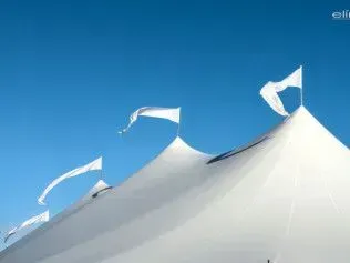 A white tent with pointed peaks topped with small white flags against a clear blue sky.