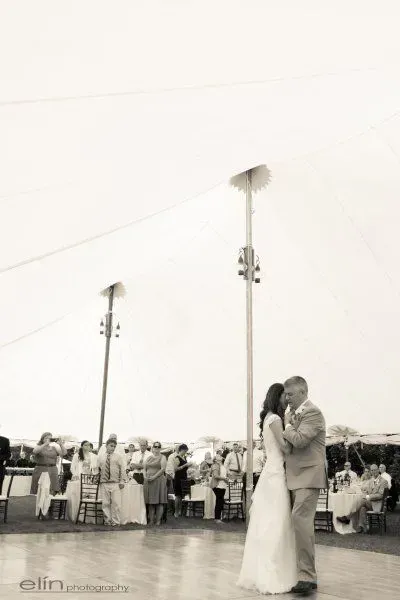 A wedding couple shares their first dance on a wooden floor inside a large tent with guests seated in the background.
