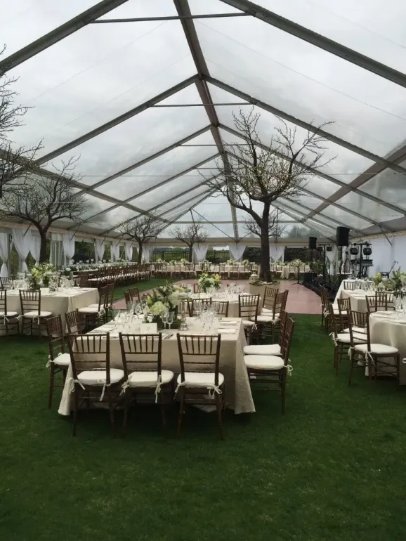 Tables with white linens and wooden chairs set up for an outdoor event under a large, clear-roofed event tent.