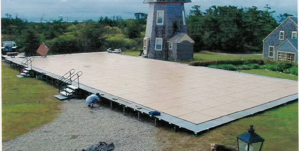 A person assembles a large, light-colored portable dance floor on a lawn in front of a windmill and a house.