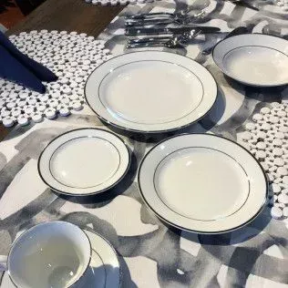 A place setting with white dinnerware featuring thin black rims, arranged on a grey and white patterned tablecloth.