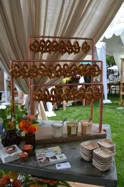 A copper stand displays rows of hanging pretzels over three jars of dipping sauce and small bowls on a stone table.