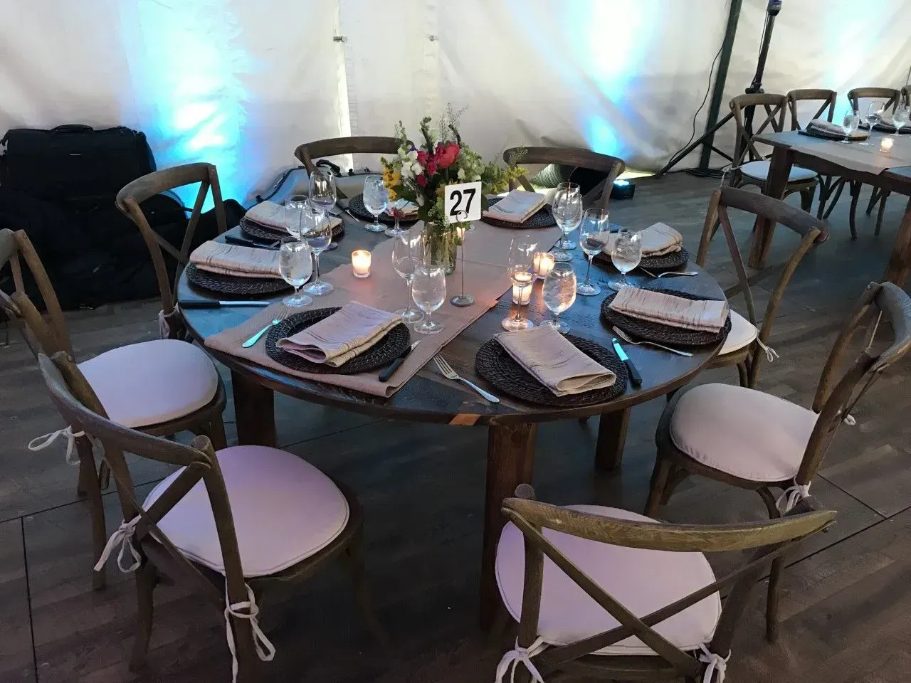 A round wedding reception table with six wooden chairs, place settings, napkins, and a floral centerpiece numbered 27.