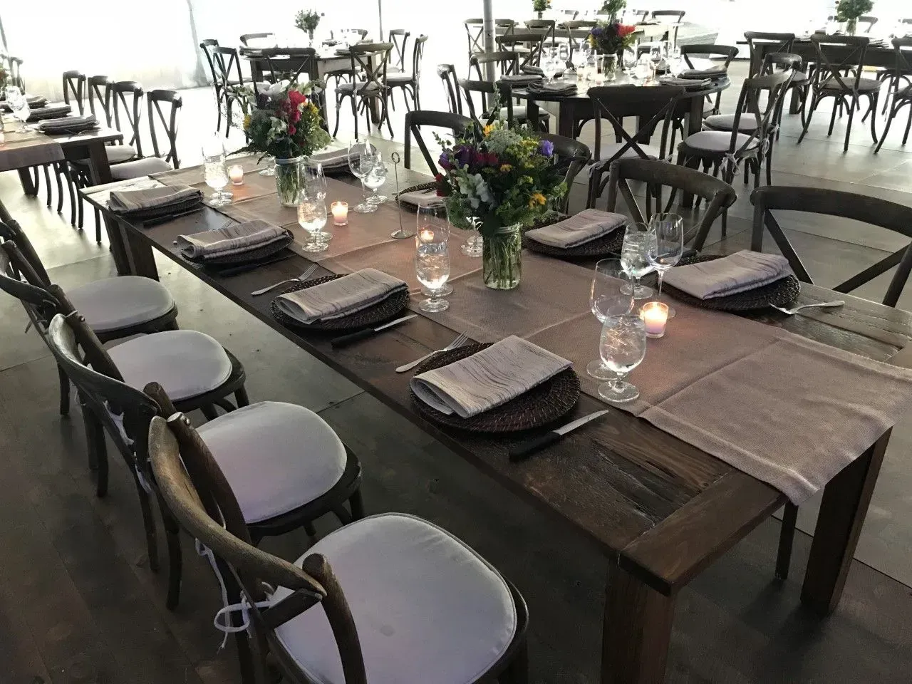 A long wooden dining table set for an event with place settings, grey linens, and centerpieces inside a white tent.