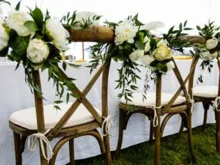 Rustic wooden cross-back chairs with cream cushions and white floral arrangements tied to the top, set for an event.