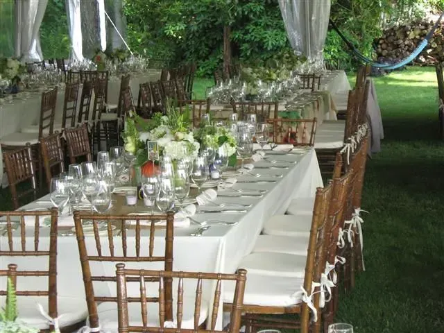 An outdoor banquet with long tables set for dinner, featuring white linens, floral centerpieces, and wooden chairs.