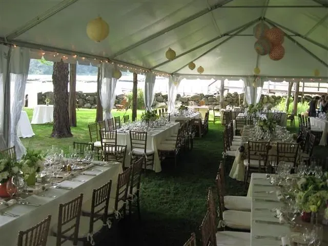 An outdoor event tent set up with long dining tables, white tablecloths, chiavari chairs, and hanging paper lanterns.