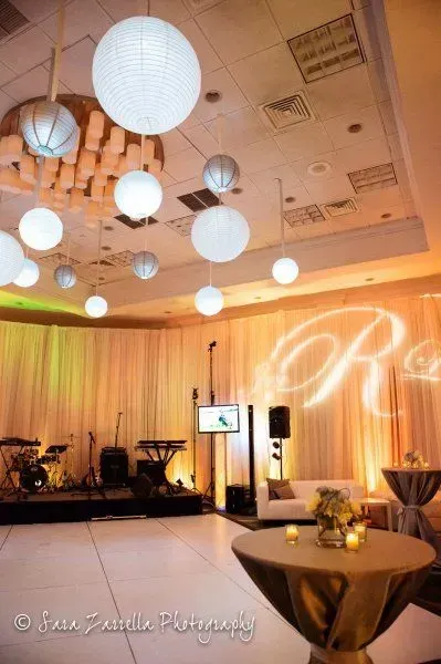 An event space with hanging globe lights, a band stage, and a monogram projection on draped walls with cocktail tables.