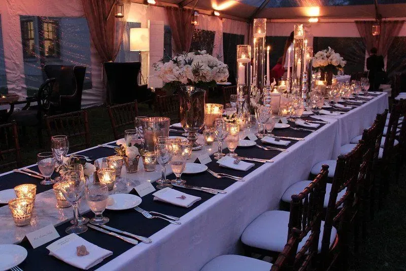 A long, elegant banquet table set for a formal event under a tent, featuring white linens, blue runners, and candles.