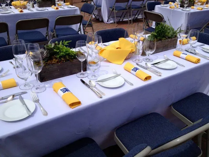 Long rectangular tables are set for an event with white tablecloths, navy chairs, yellow napkins, and greenery centerpieces.