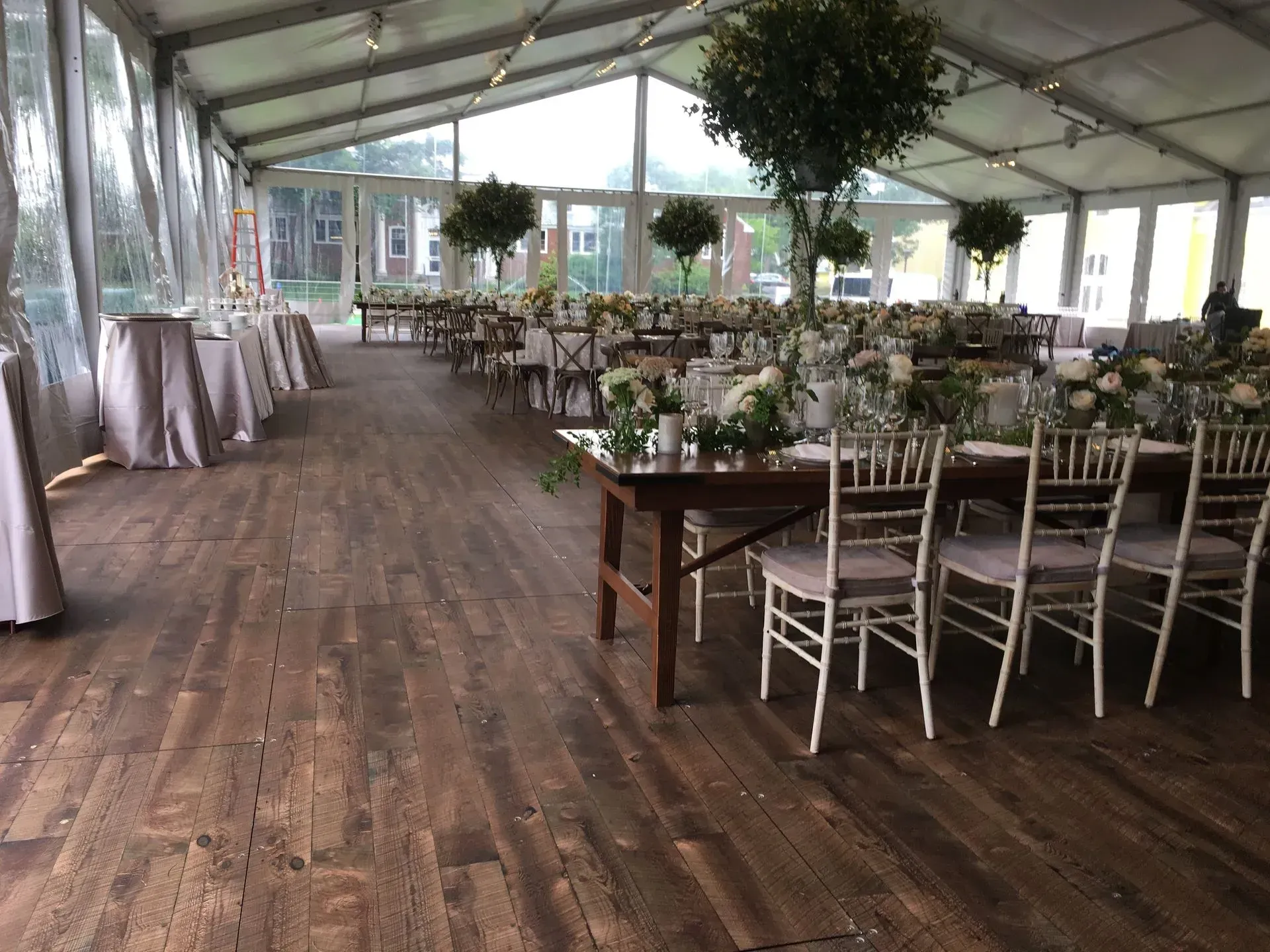 A large wedding tent featuring rows of wooden tables with white chairs and centerpieces set on a wood-plank floor.