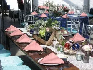 A long wooden table is set for a formal event with pink napkins, white plates, and floral centerpieces in wooden boxes.
