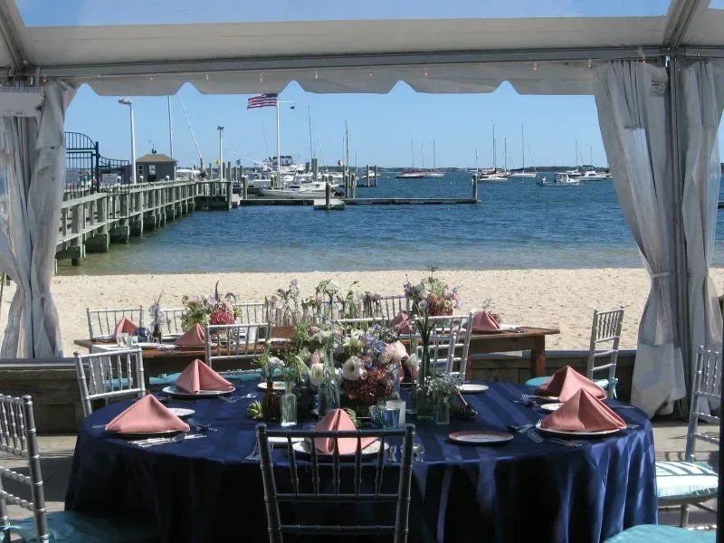 A seaside tent set with round tables, navy tablecloths, and pink napkins, overlooking a pier and boats on a sunny day.