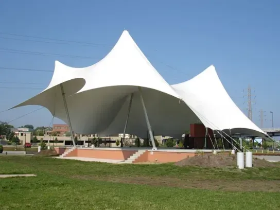 A white tensile fabric canopy structure over a raised stage platform in a grassy park setting under a clear blue sky.