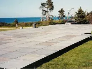 A flat stone patio overlooks a coastal landscape with trees, grass, and an ocean view under a clear sky.