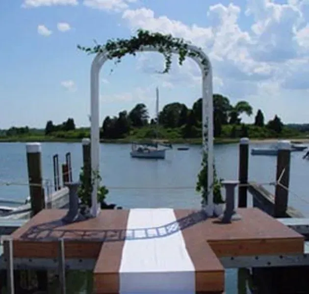 A white wedding arch adorned with greenery stands on a wooden dock over water, with a white runner and a boat in the back.