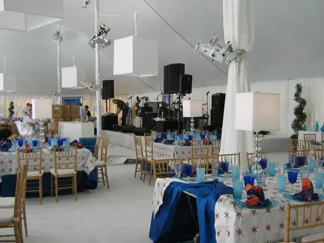Tables set for a formal event under a large white tent with blue accents, square pendant lamps, and a stage area.