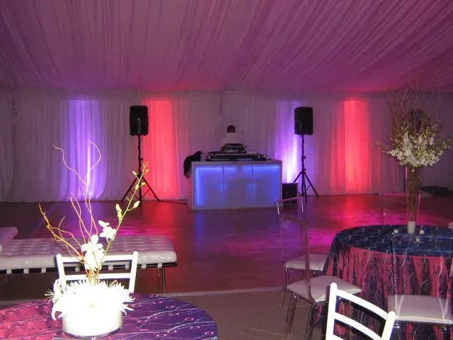 A DJ booth with purple and red accent lighting stands under a tent, surrounded by event tables and white chairs.