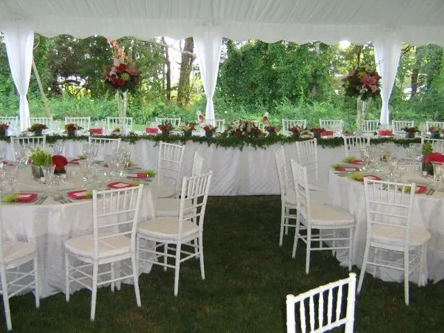Outdoor tent setup for a wedding with white chairs, round tables, and a long head table decorated with floral centerpieces.