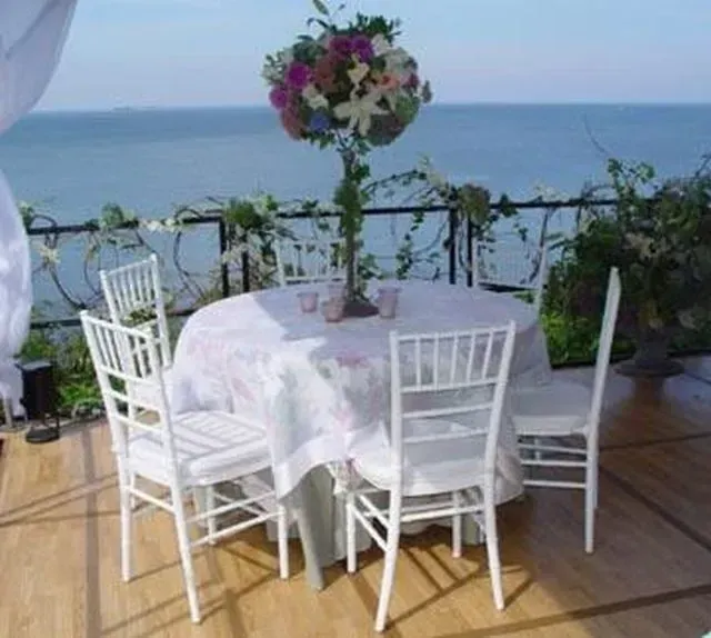 A round table with white chairs and a floral centerpiece set on a wooden deck overlooking the ocean.