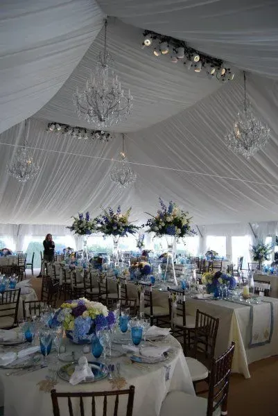 A reception tent decorated with white fabric, chandeliers, track lighting, and tables with blue floral centerpieces.