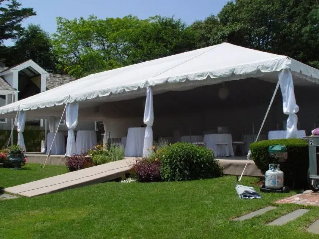 A large white event tent set up on a grassy lawn with a wooden ramp leading into it and white-covered tables inside.