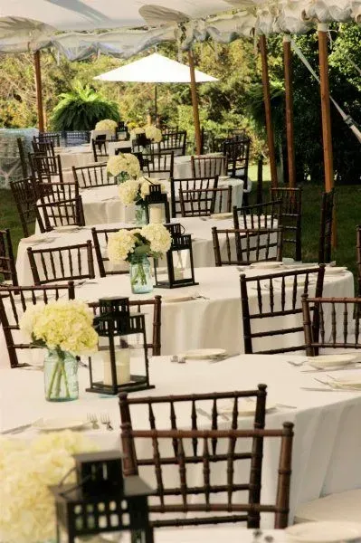 Outdoor wedding reception with rows of round tables set with white tablecloths, floral centerpieces, and dark lanterns.