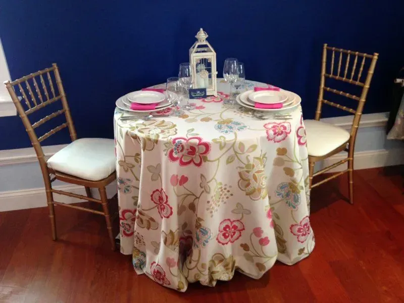 A small, round table with a floral tablecloth set for two, flanked by gold-framed chairs against a dark blue wall.