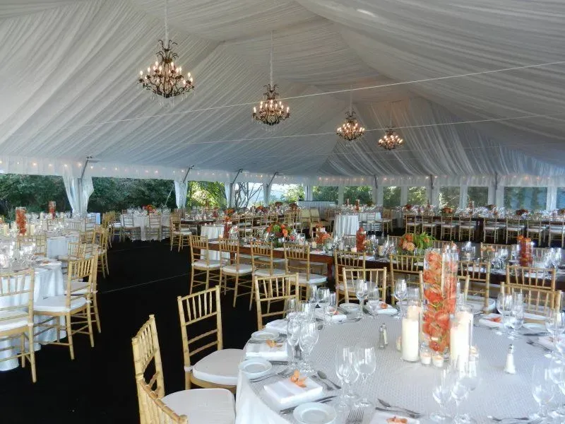An elegant event tent with draped white ceilings, crystal chandeliers, round tables with white cloths, and gold chairs.
