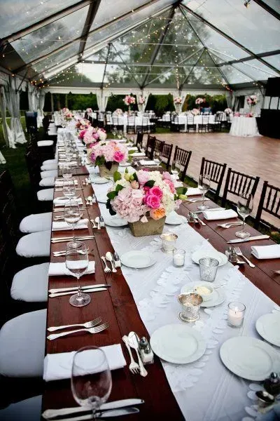 A long banquet table set for a wedding under a clear-top tent, featuring pink floral centerpieces and white place settings.