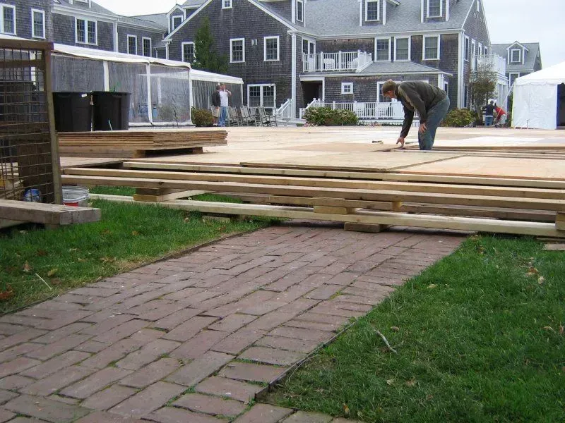 A worker installs a plywood floor over a wooden frame on a lawn in front of a shingled building and brick walkway.