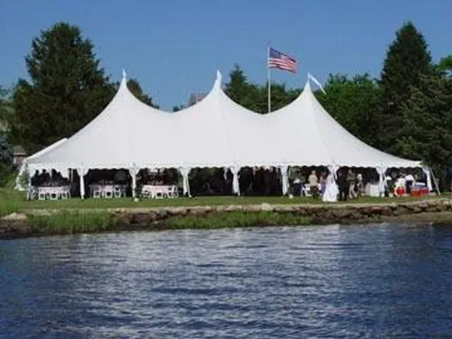 A large, white, multi-peak event tent set up on a grassy shore next to a body of water, with an American flag flying above.