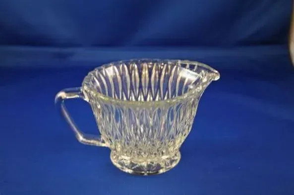 A clear glass creamer with a diamond-cut textured body and an angled handle, set against a solid blue background.