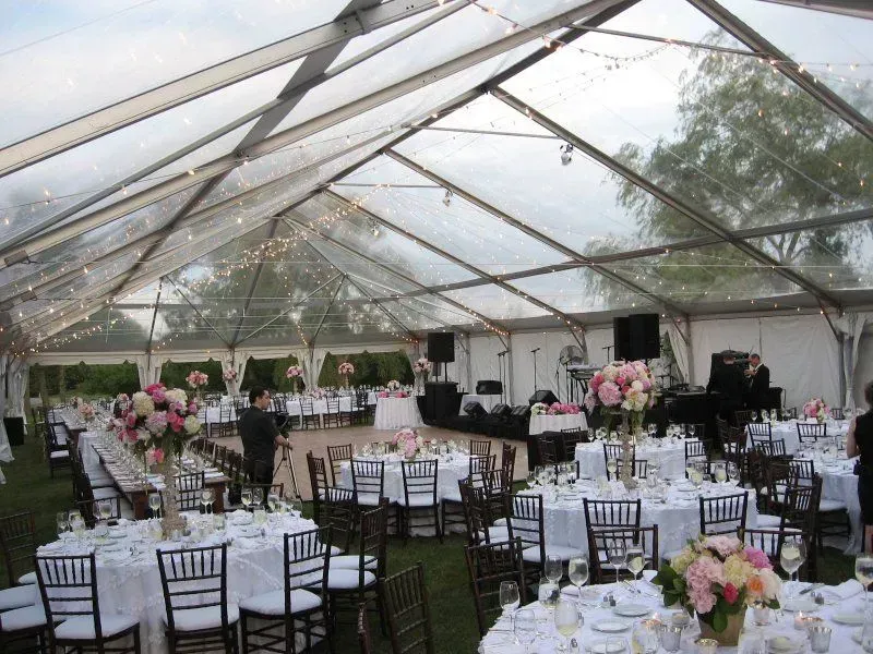 An elegant event tent with a clear ceiling, featuring round tables set for a wedding reception and a stage for music.