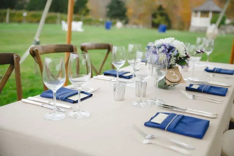 A table set for a wedding with a beige tablecloth, blue napkins, wine glasses, and a floral centerpiece with the number 3.