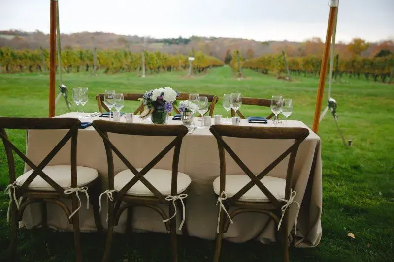 A table set for dining under a tent, featuring wooden cross-back chairs facing a scenic vineyard.