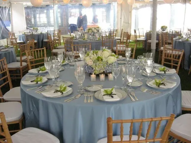 Round tables with blue tablecloths and gold chairs set for a formal event in a tent, with floral centerpieces.