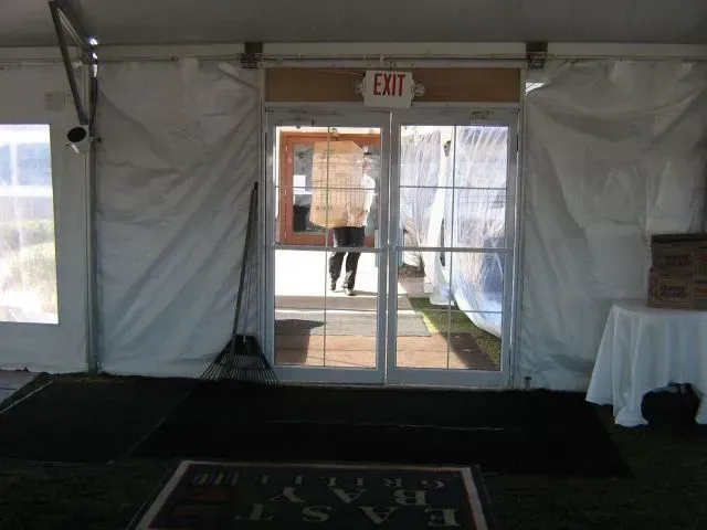 A view through glass double doors of a tent looking out to a parking area where a person carries a large cardboard box.