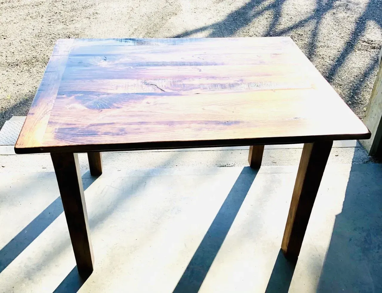 A rustic, rectangular wooden dining table with dark brown legs stands on a concrete floor in natural sunlight.