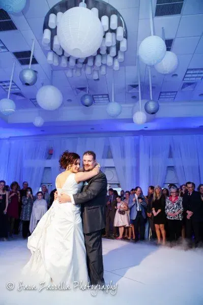 A newlywed couple dances in the center of a brightly lit event space decorated with hanging globes and sheer curtains.