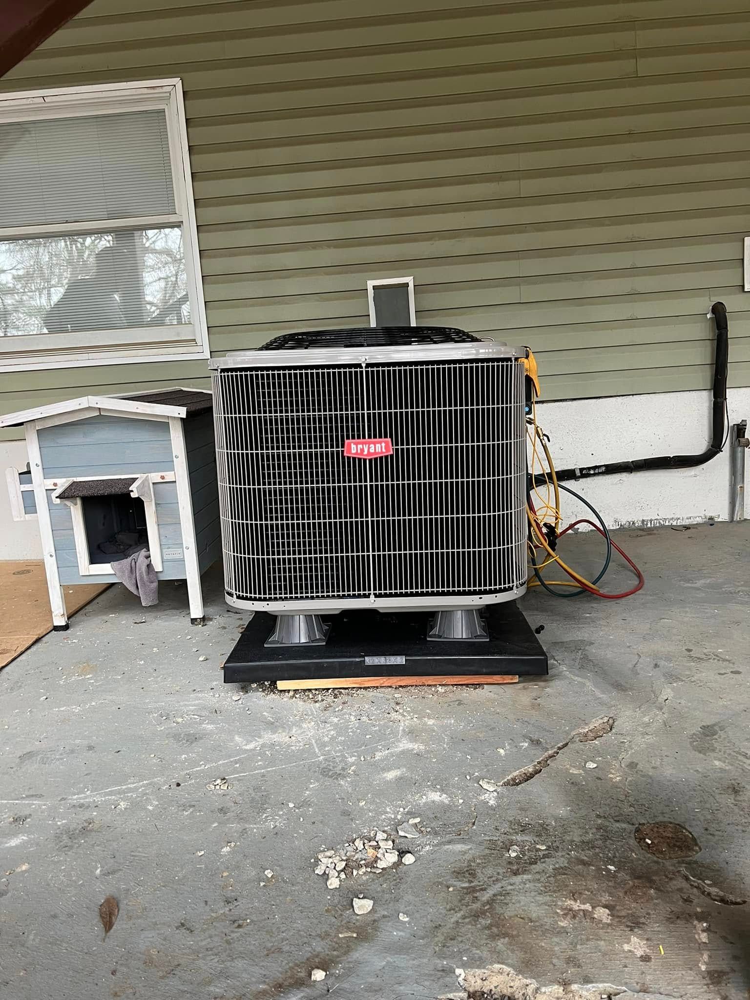 An outdoor air conditioning unit sits on a black mounting base against a house with light green horizontal siding.