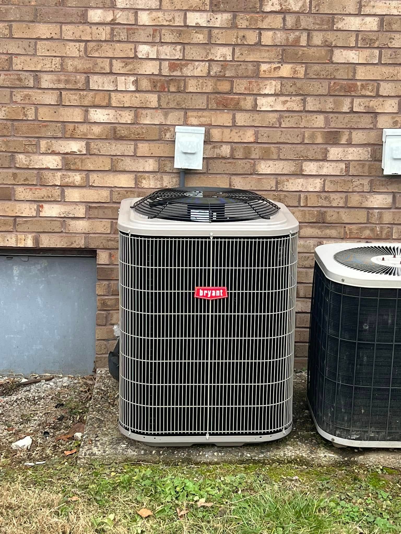 A beige HVAC outdoor unit stands on a concrete pad against a brick wall, positioned next to a partially visible unit.