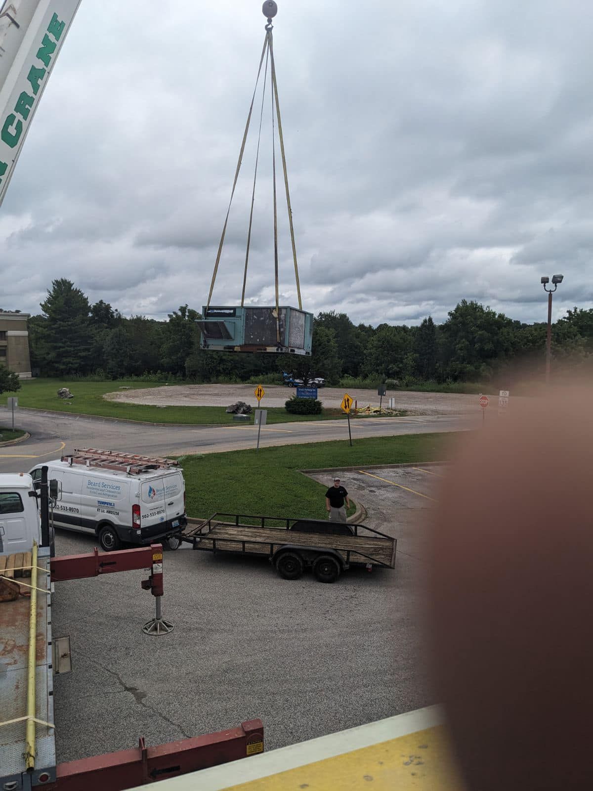 A crane lifts a large container into the air above a parking lot with a work van and a trailer nearby.
