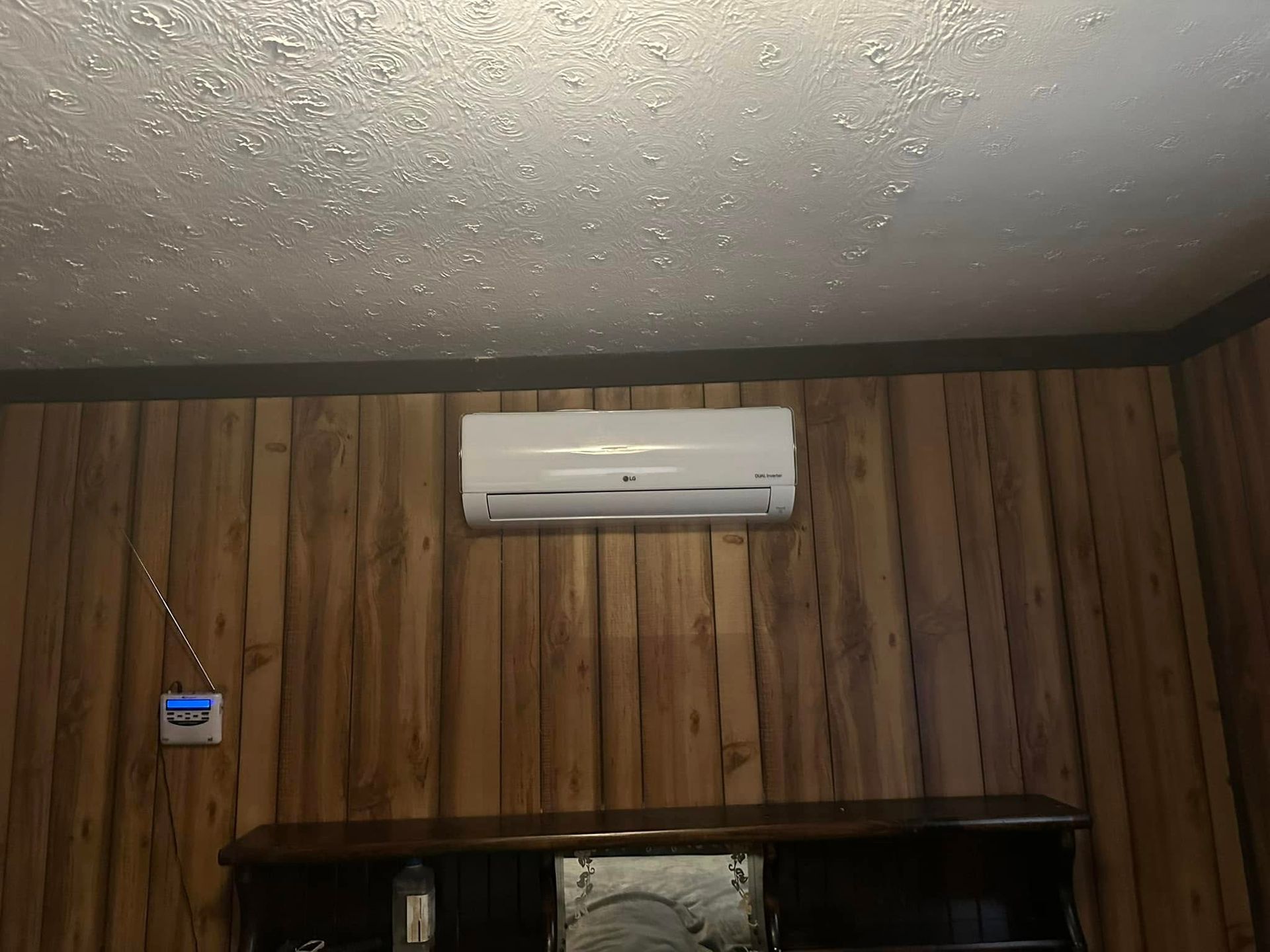 A white wall-mounted air conditioner unit centered on a brown wood-paneled wall below a textured white ceiling.
