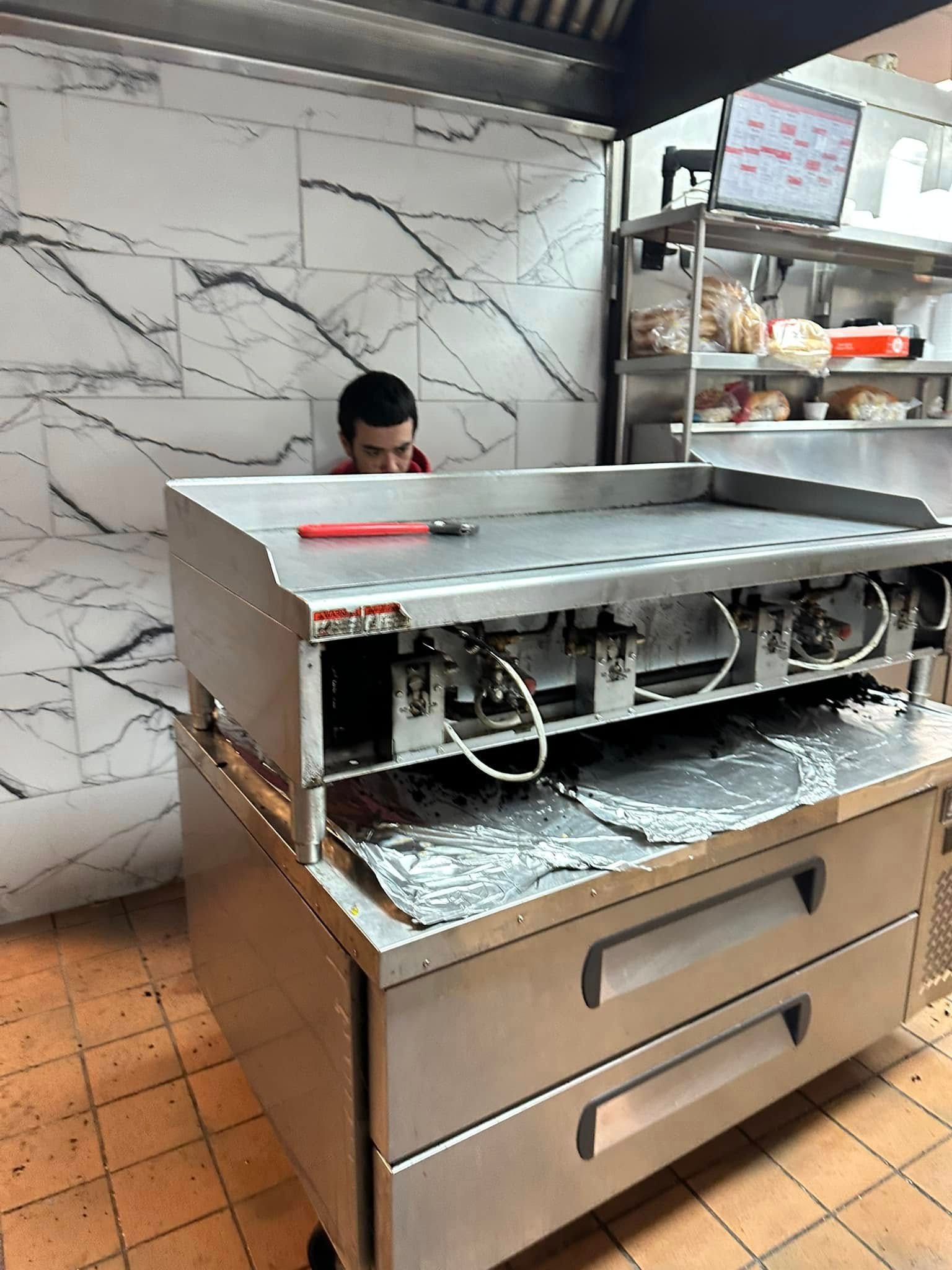 A kitchen worker stands behind a stainless steel commercial flat-top grill in a tiled room.
