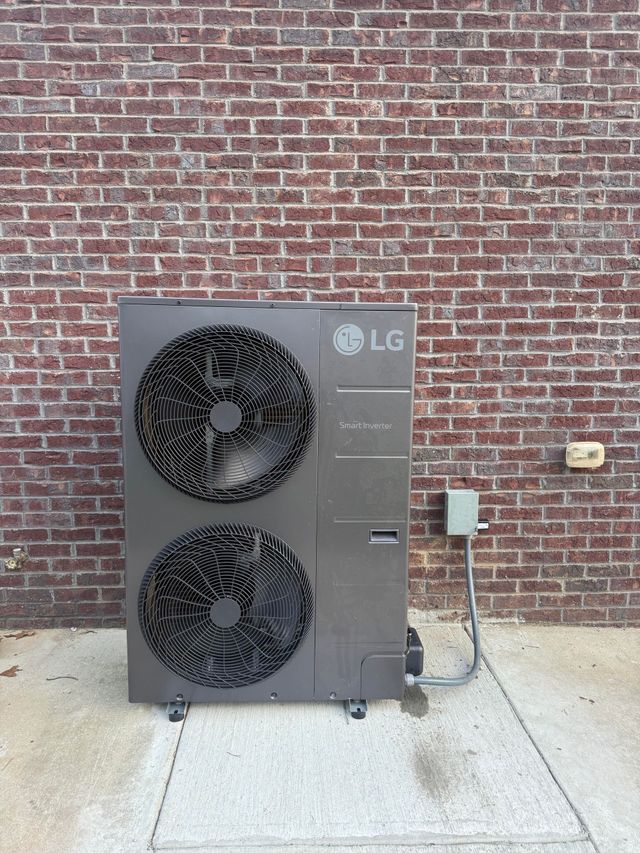 A beige HVAC outdoor unit stands on a concrete pad against a brick wall, positioned next to a partially visible unit.