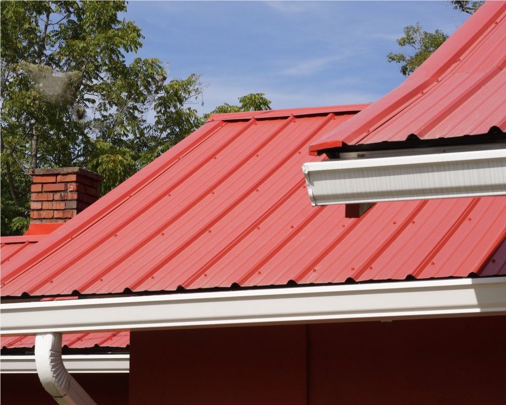 Red metal roof with white trim and a brick chimney under a blue sky, with trees nearby